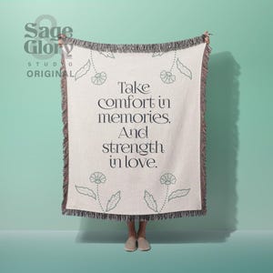 May include: A cream-colored woven throw blanket featuring dark green floral illustrations and the text "Take comfort in memories, And strength in love." The blanket has a fringed edge and is displayed vertically with a dark border.