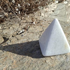Könnte beinhalten: Ein weißer, glatter, pyramidenförmiger Kristall auf einer rauen, grauen, strukturierten Oberfläche.