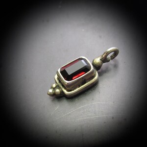 May include: A silver pendant with a rectangular, deep red gemstone. The pendant has a decorative design with a loop at the top for a chain. The gemstone is set in a silver frame, and the pendant has an antique look.