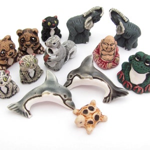 May include: A collection of small, hand-painted animal figurines. The assortment includes a variety of creatures such as cats, bears, elephants, a squirrel, a dolphin, a turtle, a frog, and a Buddha statue. The figurines are in various colors, including gray, brown, and green.