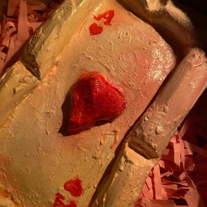 以下が含まれることがあります： 手作りのトランプ、ハートのエースが箱に入っています。カードはベージュとピンクの色合いで塗装されており、赤いハートのシンボルと文字「A」が描かれています。カードはピンク色の紙のストリップで囲まれています。