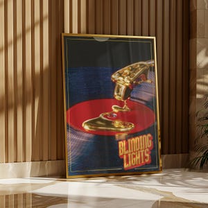 Puede incluir: Una impresión artística enmarcada que presenta una aguja de tocadiscos dorada goteando sobre un disco de vinilo rojo. El fondo es azul oscuro con las palabras "BLINDING LIGHTS" en rojo y dorado. El marco es dorado.