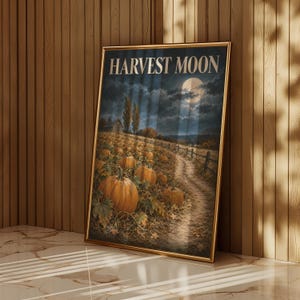 May include: A rectangular framed wall art print featuring a nighttime landscape with a pumpkin patch, a dirt path, and a full moon in the sky. The text HARVEST MOON is printed in large, light-colored capital letters at the top of the artwork.