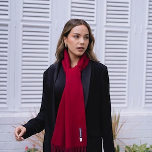 Op de afbeelding: Een vrouw draagt een zwarte blazer over een zwart topje en een blauwe jeans. Ze draagt een rode sjaal met franjes om haar nek.