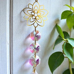 May include: A gold-coloured metal suncatcher featuring a floral design, connected to three iridescent crystal beads and a teardrop-shaped crystal. The text "PrismBeads" is visible.
