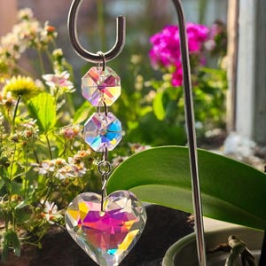 May include: A decorative hanging prism featuring a heart-shaped crystal pendant and two octagonal crystals. The crystals display iridescent colours. The prism hangs from a silver metal hook. The text "PrismBeads" is visible at the bottom.