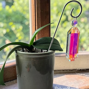 May include: An orchid plant in a grey pot sits on a wooden windowsill. A decorative prism suncatcher hangs from a metal hook. The prism is a multi-coloured crystal with a faceted, rectangular shape. The text "PrismBeads" is visible.