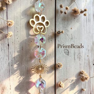 May include: A hanging suncatcher ornament featuring a gold-toned metal paw print charm with a central heart cutout, a sunburst charm, and multiple iridescent faceted crystal beads in various shapes, including a teardrop, suspended on a gold chain.