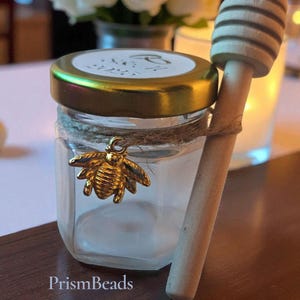 May include: A small, clear glass jar with a gold lid and a gold bee charm tied with twine. A wooden honey dipper rests beside the jar. The jar has a white label on the lid. The text "PrismBeads" is at the bottom of the image.