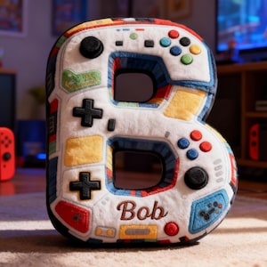 Puede incluir: Un cojín de felpa blanco con forma de letra "B" con detalles coloridos con temática de mando. El cojín presenta una cruceta, botones y el nombre "Bob" bordado en marrón. El diseño incorpora detalles en rojo, azul, amarillo y verde.