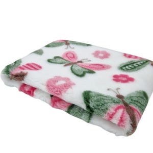 May include: Folded white plush blanket with a butterfly and floral pattern. The butterflies have green wings and pink accents. Pink flowers and green leaves are also scattered across the blanket.