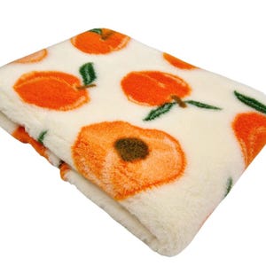 May include: Folded plush blanket with a repeating pattern of orange peaches and green leaves on a cream background. The blanket has a soft, fuzzy texture, ideal for adding warmth and comfort to any space.