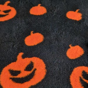 May include: Black Halloween rug with orange pumpkin designs. The rug features a repeating pattern of smiling jack-o'-lantern faces and smaller pumpkin shapes. The rug has a soft, textured appearance, ideal for seasonal decor.
