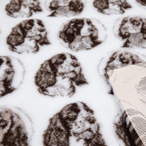 May include: White fabric with a repeating pattern of brown dog faces. The fabric has a soft, fuzzy texture.