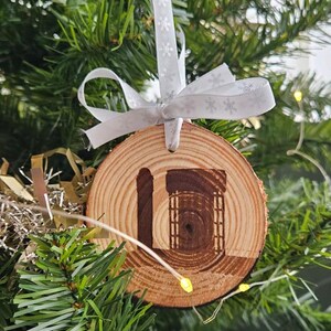 May include: A round wooden Christmas tree decoration with a natural wood grain. The ornament has a carved design of a building and the letter "L". It is hung by a white ribbon with snowflake patterns.