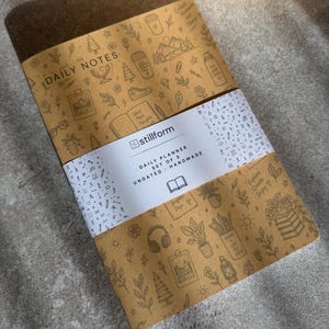 May include: A set of three tan daily planners with a white paper band featuring the text stillform, DAILY PLANNER, SET OF 3, UNDATED, HANDMADE. The covers are decorated with brown line art illustrations of plants, mountains, coffee cups, and books.