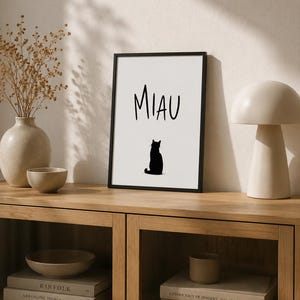 Könnte beinhalten: Ein rechteckiger, schwarz gerahmter Kunstdruck mit weißem Hintergrund, auf dem das Wort MIAU in schwarzen Großbuchstaben über der Silhouette einer schwarzen Katze steht, die sitzt und vom Betrachter wegblickt, mittig platziert.