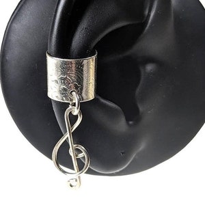 May include: Silver ear cuff with a treble clef charm hanging from it. The cuff is decorated with a floral pattern.
