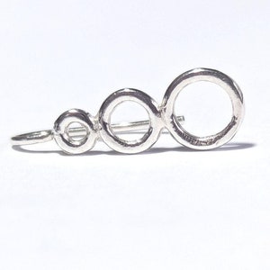 May include: Silver ear cuff with three connected circles of varying sizes. The circles are smooth and have a slightly textured finish.