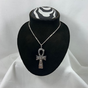 May include: A silver ankh pendant necklace on a black display stand with a zebra print top. The ankh is a symbol of life in ancient Egyptian culture.