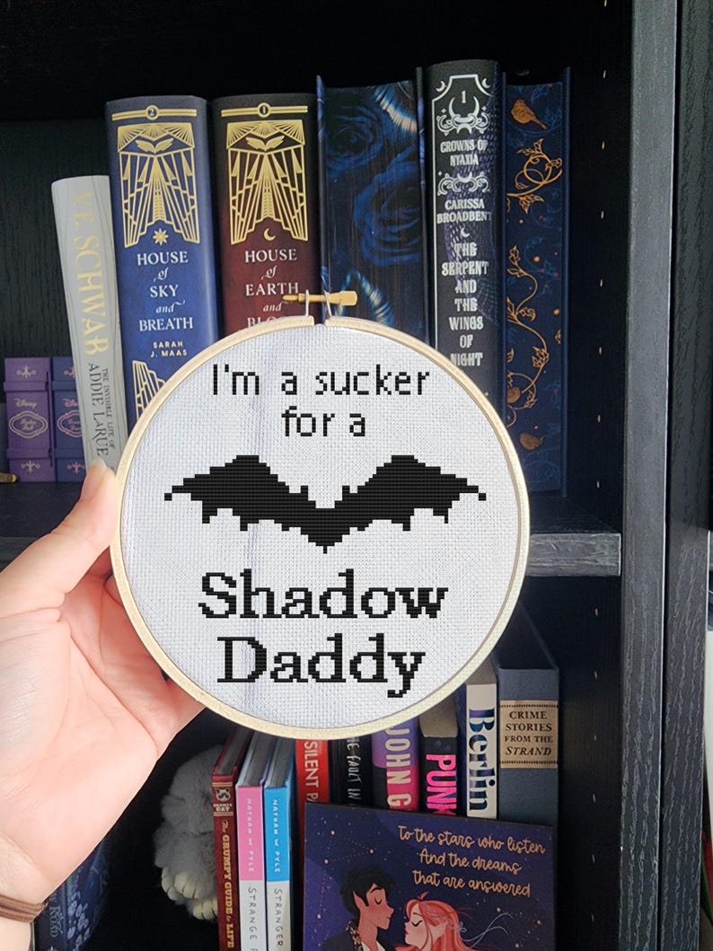 Hand holding a finished cross stitch embroidery hoop displaying I'm a Sucker for a Shadow Daddy pattern with bat design, photographed in front of a romantasy bookshelf featuring House of Earth and Blood and House of Sky and Breath by Sarah J Maas