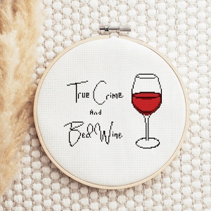 Op de afbeelding: Een witte borduurring met de tekst "True Crime and Red Wine" en een rood wijnglas met rode vloeistof.