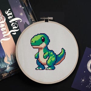 May include: A green and blue pixelated dinosaur cross stitch pattern on a white canvas in a wooden embroidery hoop.