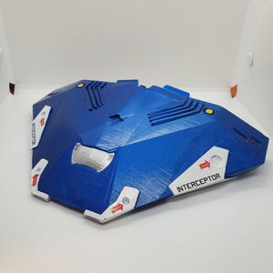 Op de afbeelding: Een blauw en wit 3D-geprint model van een ruimteschip met het woord "INTERCEPTOR" erop gedrukt.