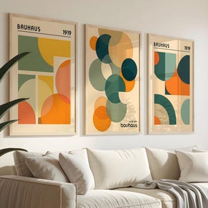 Könnte beinhalten: Drei gerahmte Bauhaus-Kunstdrucke mit geometrischen Designs in Orange-, Grün- und Gelbtönen. Die Drucke werden über einem cremefarbenen Sofa mit einer grauen Decke präsentiert. Links steht eine Topfpflanze.