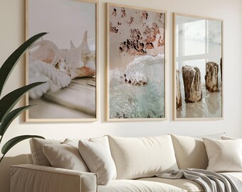 May include: A living room scene featuring a beige sofa with throw pillows and a gray blanket. Above the sofa are three framed art prints with beach-themed images. A large potted plant is on the left, and a window with a small plant is on the right.
