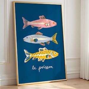 Op de afbeelding: Een ingelijste kunstdruk met drie gestileerde vissen in roze, blauw en geel tegen een diepblauwe achtergrond. Het kunstwerk zit in een gouden lijst en bevat de Franse zin "le poisson" in witte letters.