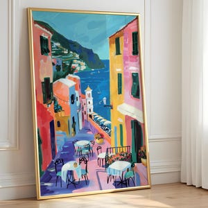 May include: A framed art print showing a vibrant street scene with buildings in red, pink, orange, and blue. The artwork depicts a coastal town with tables and chairs on the street, overlooking the sea and mountains in the background.