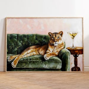 May include: A framed artwork shows a lioness lounging on a green velvet sofa. A martini with olives is on a small, round table. The background is a soft pink, and the frame is light wood.