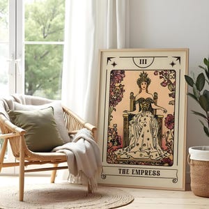 May include: A framed tarot card print of "The Empress" in a light wood frame. The artwork features a seated woman in a gown and crown, surrounded by floral elements. The print is displayed next to a woven chair and a window.