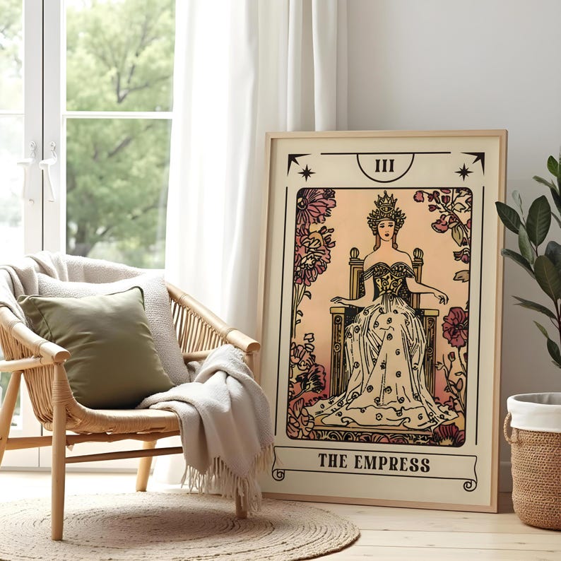 May include: A framed tarot card print of "The Empress" in a light wood frame. The artwork features a seated woman in a gown and crown, surrounded by floral elements. The print is displayed next to a woven chair and a window.
