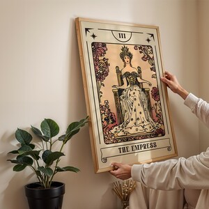 May include: A framed tarot card print of "The Empress" in a vintage style. The artwork features a woman in a gown and crown seated on a throne, surrounded by floral elements. The print has a beige background with black and pink accents.