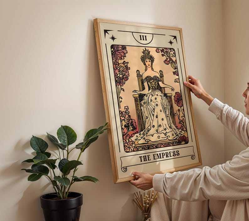 May include: A framed tarot card print of "The Empress" in a vintage style. The artwork features a woman in a gown and crown seated on a throne, surrounded by floral elements. The print has a beige background with black and pink accents.