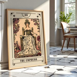 May include: A framed print of The Empress tarot card. The artwork features a woman in a gown and crown seated on a throne, surrounded by floral elements. The color palette includes beige, pink, and black, with the title "THE EMPRESS" at the bottom.