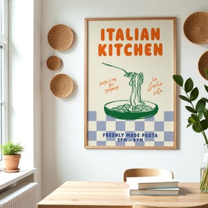 May include: A framed poster with a white background and a blue and white checkered pattern at the bottom. The poster features a green illustration of a bowl of pasta with a fork and text that reads "ITALIAN KITCHEN", "Pasta is my love language", "La Dolce Vita", and "FRESHLY MADE PASTA 3PM-9PM".