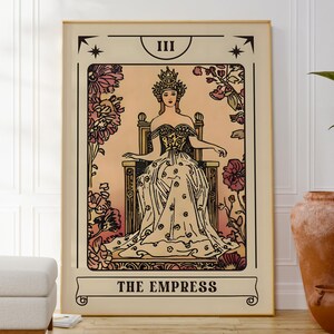 May include: A framed tarot card print featuring "The Empress." The artwork depicts a woman in a crown and gown seated on a throne, surrounded by floral elements. The color palette includes beige, pink, and black, with the title at the bottom.