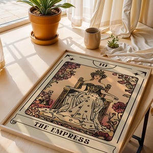 May include: A framed tarot card print of "The Empress" in a light wood frame. The artwork features a seated figure with a crown, surrounded by floral elements in shades of pink and cream. The text "THE EMPRESS" is at the bottom.
