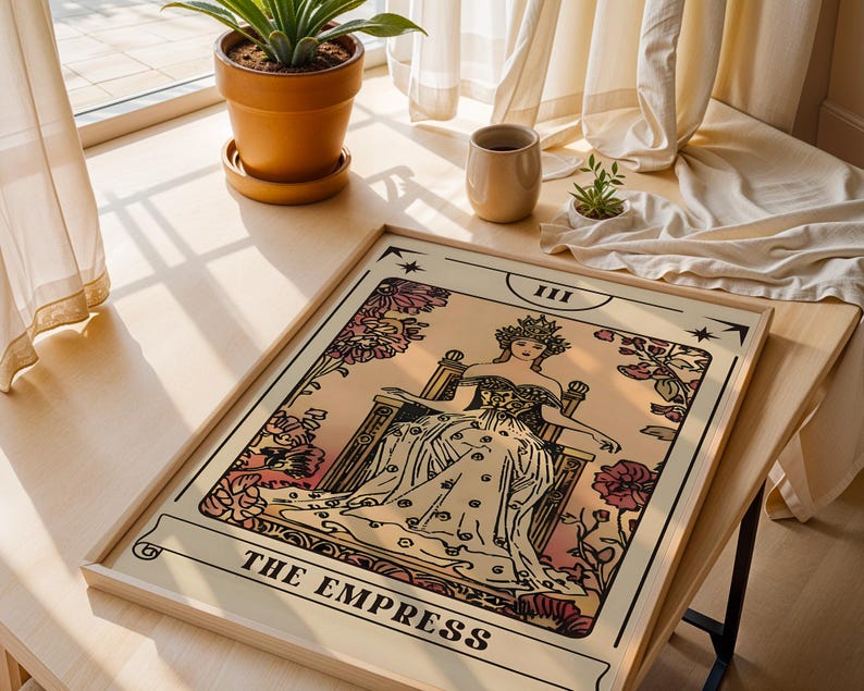 May include: A framed tarot card print of "The Empress" in a light wood frame. The artwork features a seated figure with a crown, surrounded by floral elements in shades of pink and cream. The text "THE EMPRESS" is at the bottom.