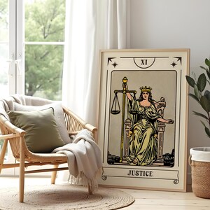 May include: A framed tarot card print featuring the Justice card. The artwork depicts a robed figure holding scales, with the word "JUSTICE" at the bottom. The print is displayed next to a wicker chair with a green pillow and a beige blanket.