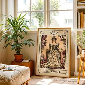 May include: A framed tarot card print of "The Empress" in a room with plants and a window. The artwork features a regal woman in a gown and crown, surrounded by floral designs. The print is displayed in a light-colored wooden frame.