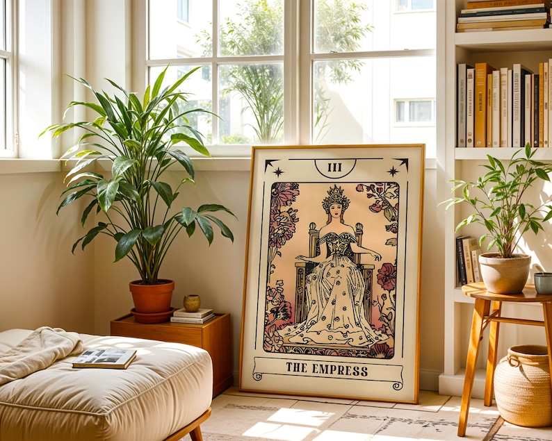 May include: A framed tarot card print of "The Empress" in a room with plants and a window. The artwork features a regal woman in a gown and crown, surrounded by floral designs. The print is displayed in a light-colored wooden frame.