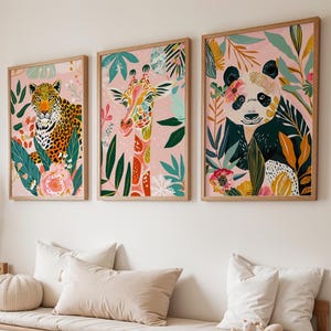 May include: Set of three framed art prints featuring colorful animal illustrations. The prints depict a leopard, giraffe, and panda bear, each surrounded by vibrant floral and foliage elements against a pink background. The frames are made of light-colored wood.