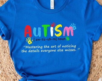 May include: A blue t-shirt with the word "AUTISM" written in colorful letters. The word is defined as "[aw-tiz-uh-m] noun" and the definition continues with "Mastering the art of noticing the details everyone else misses."