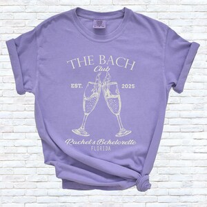 Pu&ograve; includere: Una maglietta lilla con le maniche arrotolate. Sul davanti &egrave; presente la scritta "THE BACH Club" sopra due calici di champagne che si toccano. Sotto, le parole "Rachel's Bachelorette FLORIDA" e "EST. 2025".