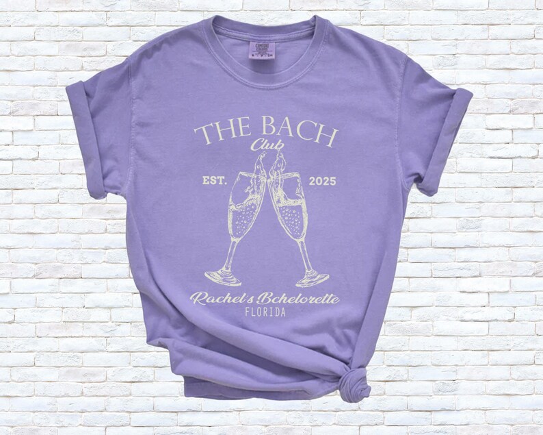 Pu&ograve; includere: Una maglietta lilla con le maniche arrotolate. Sul davanti &egrave; presente la scritta "THE BACH Club" sopra due calici di champagne che si toccano. Sotto, le parole "Rachel's Bachelorette FLORIDA" e "EST. 2025".