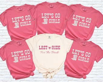 May include: A collection of pink t-shirts with the text "LET'S GO GIRLS" and a white cowboy boot graphic. One cream-colored shirt reads "LAST RIDE For The Bride" in maroon. The shirts are short-sleeved.